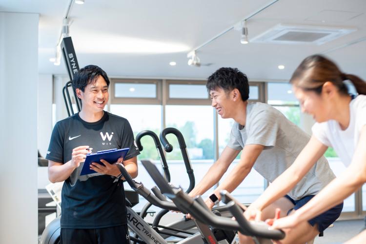 バイクを漕ぐクライアントと状態確認をするアスレティックトレーナー